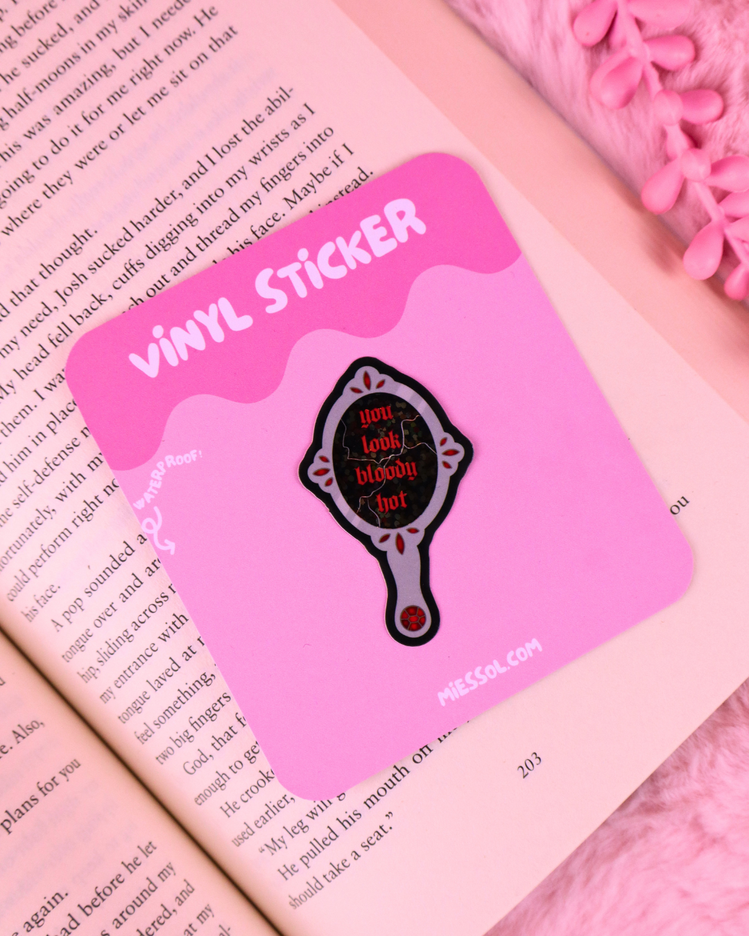 A pink vinyl sticker featuring a hand mirror design with the text 'You look bloody hot' in red, placed on an open book with a pink fuzzy blanket in the background.