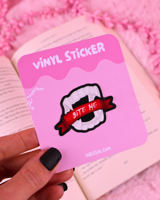 A hand with black nail polish holds a pink card displaying a vinyl sticker. The sticker shows a white mouth with teeth and a red banner reading 'BITE ME'.