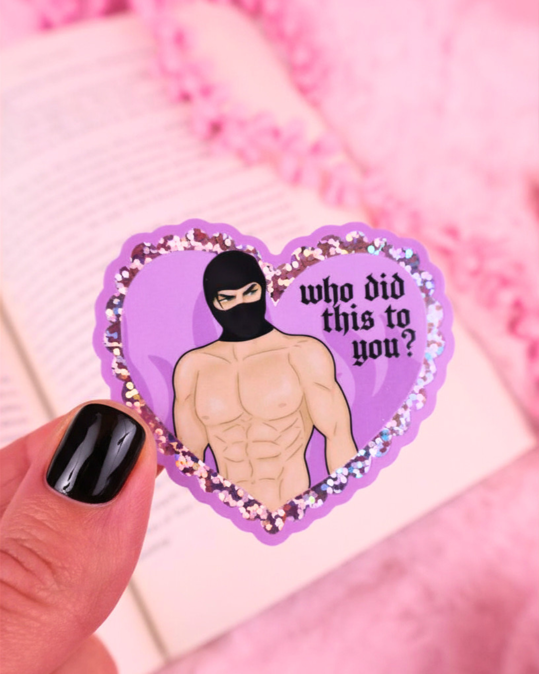 Heart-shaped sticker with a person wearing a black mask and text 'who did this to you?' held by a hand against a pink background. booktok masked men, fictional men, dark romance