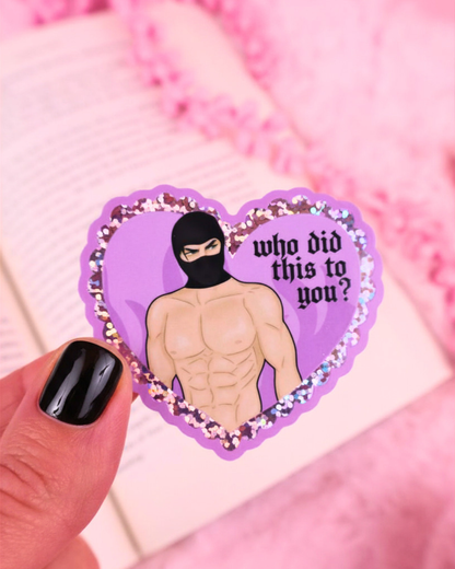 Heart-shaped sticker with a person wearing a black mask and text 'who did this to you?' held by a hand against a pink background. booktok masked men, fictional men, dark romance
