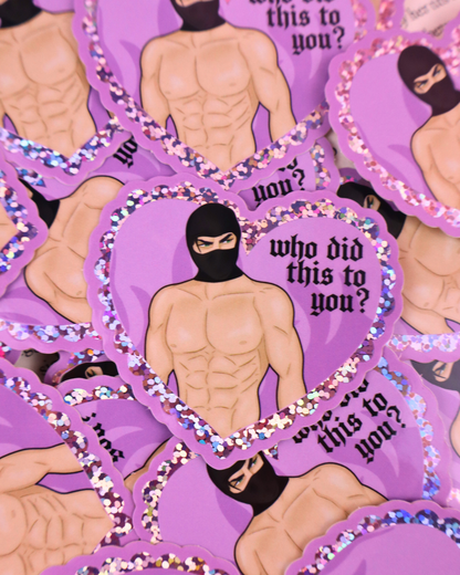 Heart-shaped sticker with a person wearing a black mask and text 'who did this to you?' held by a hand against a pink background. booktok masked men, fictional men, dark romance